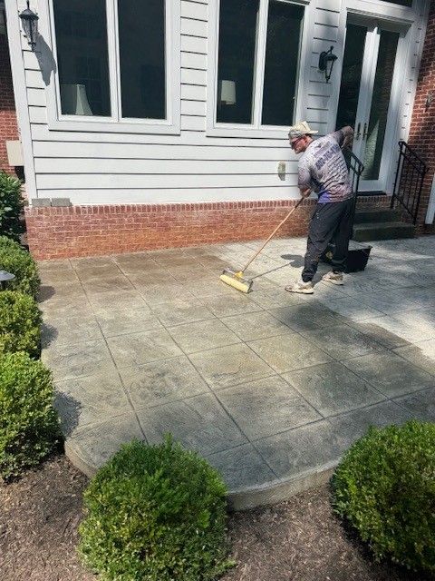 Person using a brush to clean a concrete patio next to a house with windows and shrubbery.