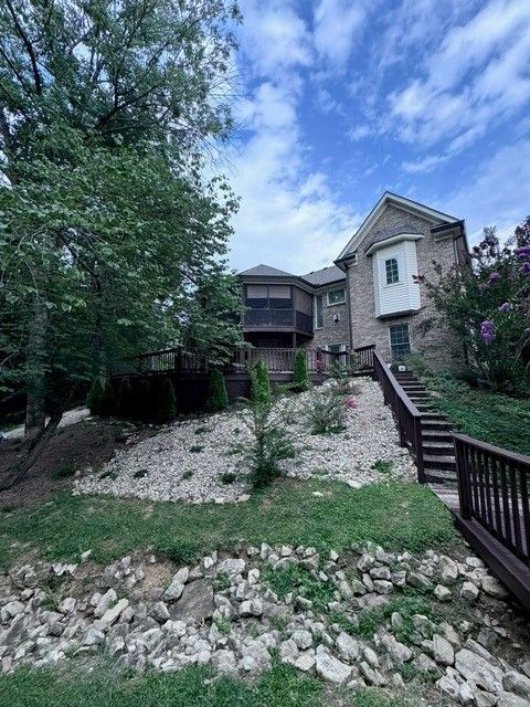 House with a brick exterior, wooden deck, and steps on a grassy hill, under a cloudy blue sky.
