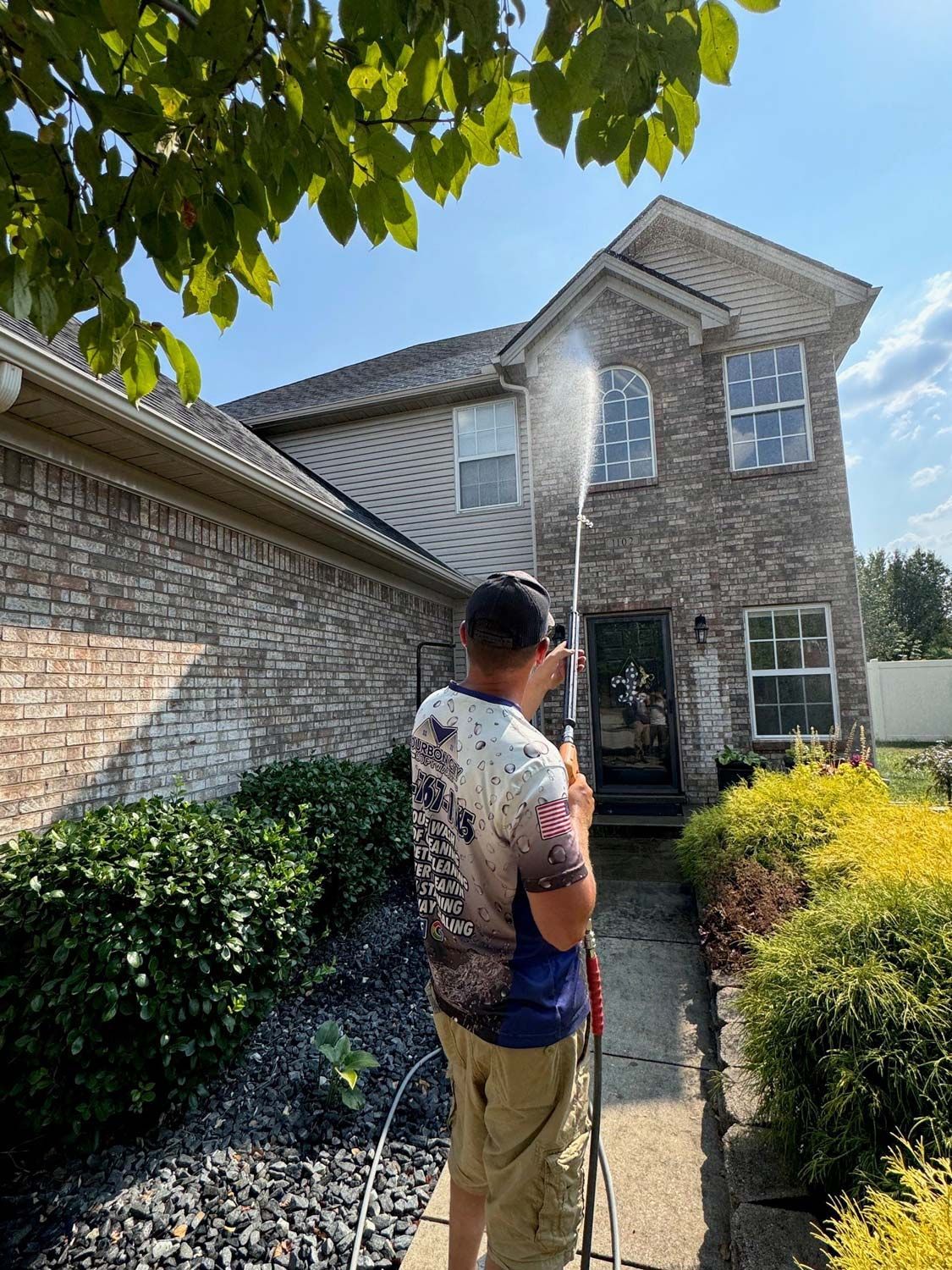 Man power washing a two-story brick house on a sunny day. He's holding a hose, spraying water.