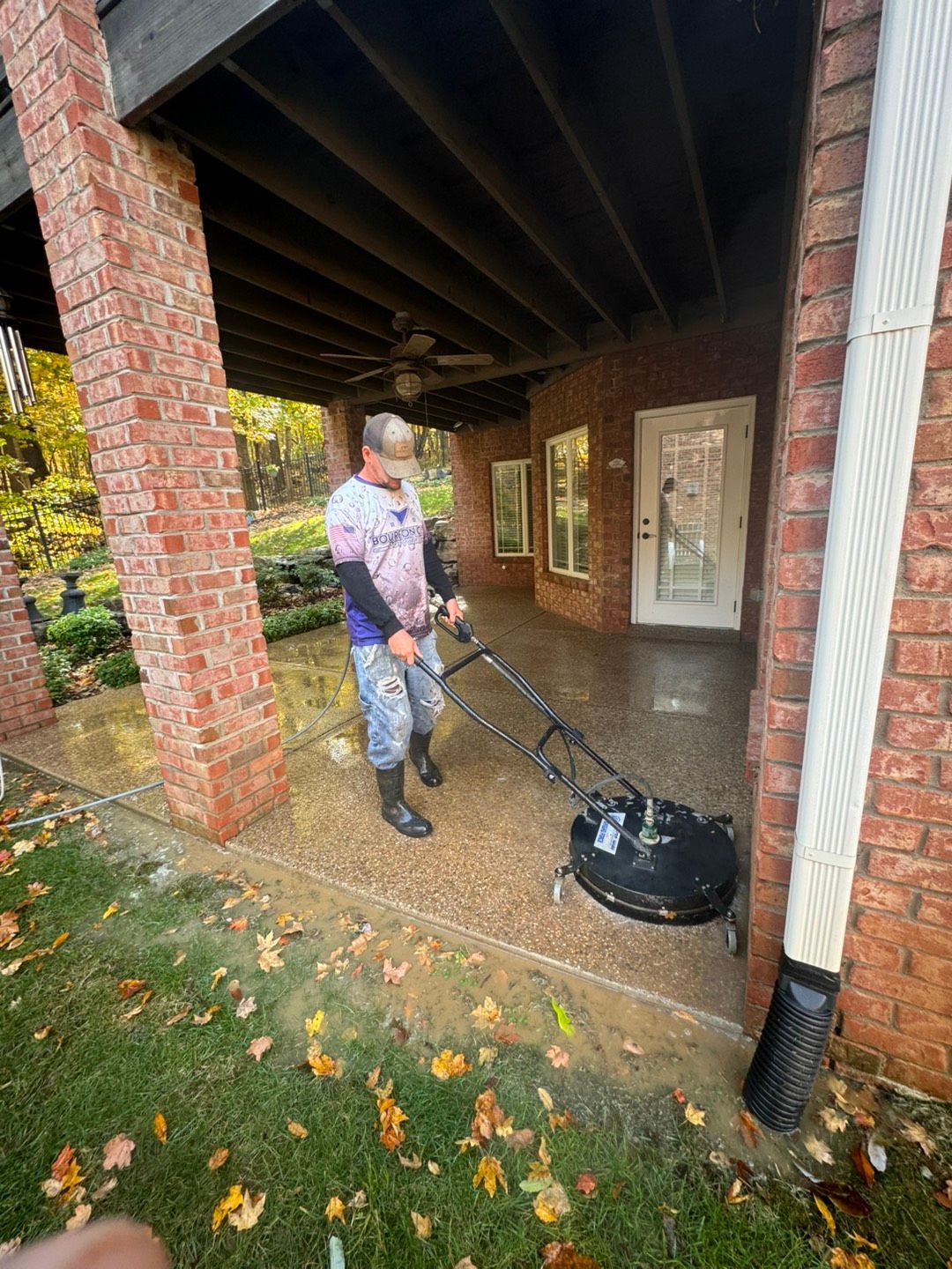 A person pressure washes a concrete patio under a brick-columned porch; fall leaves scattered.