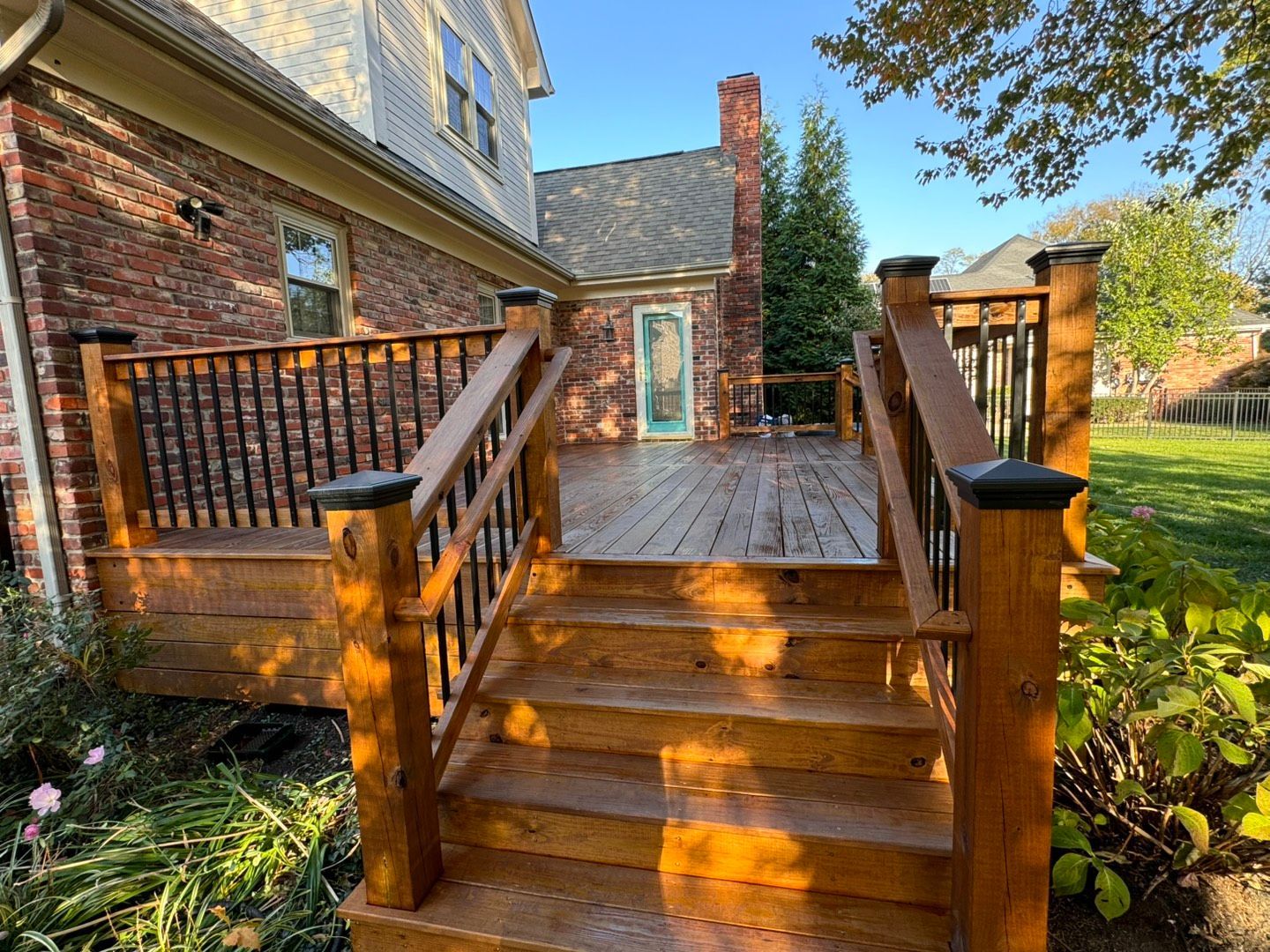 Wooden deck with stairs, attached to a brick house. Sunny day with green lawn and small building in the background.