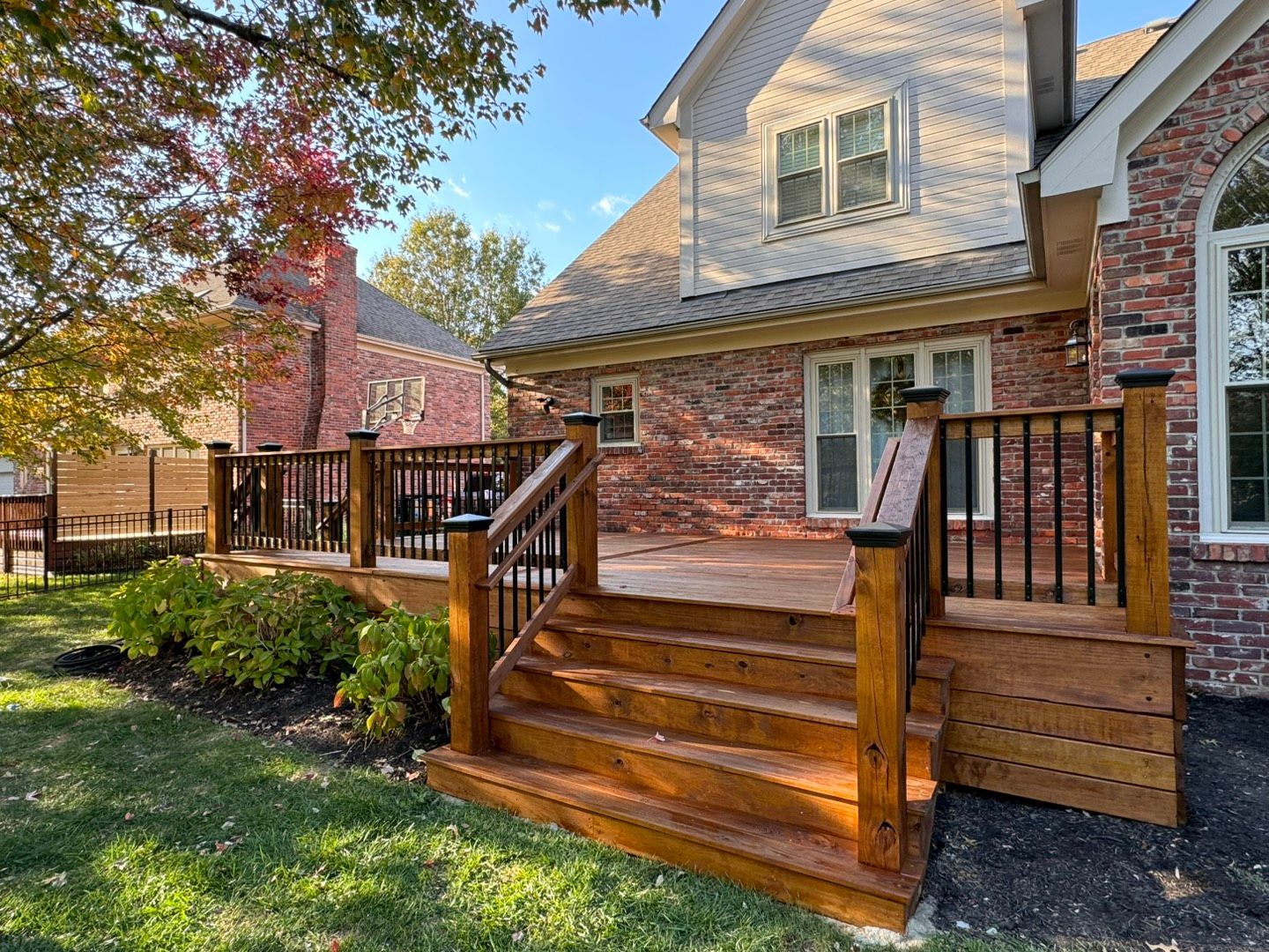 Wooden deck with stairs attached to a red brick house. Railing is dark brown, surrounding lush yard.