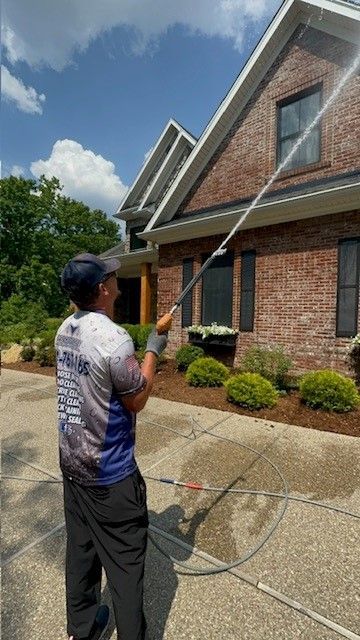Man pressure washing the exterior of a brick house with a long wand under a bright blue sky.