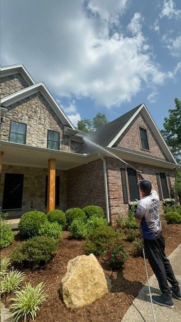 Man pressure washing a house with stone and brick exterior under a cloudy sky.