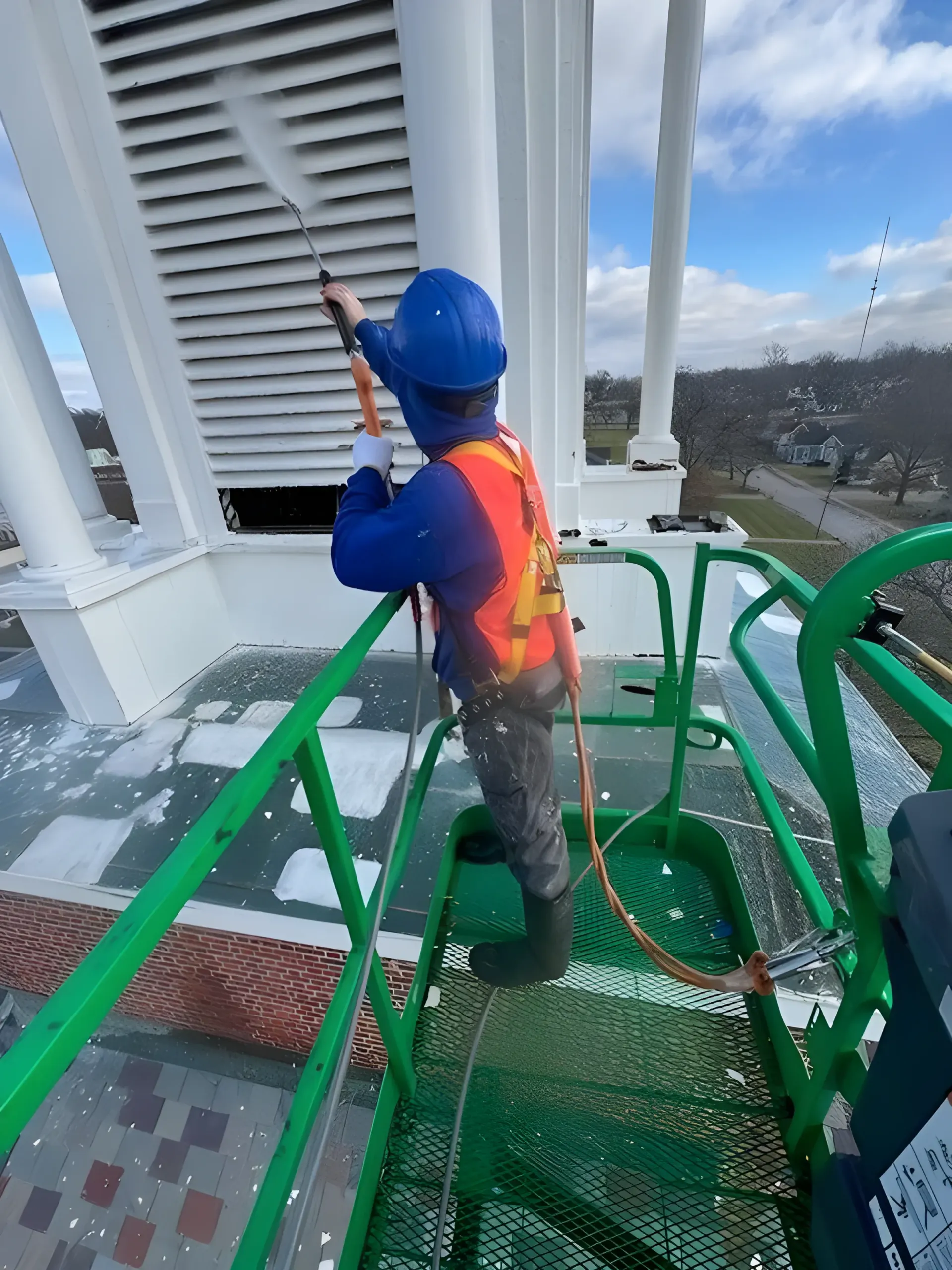 Person in safety gear power washing white building vent from a green platform, blue sky in background.