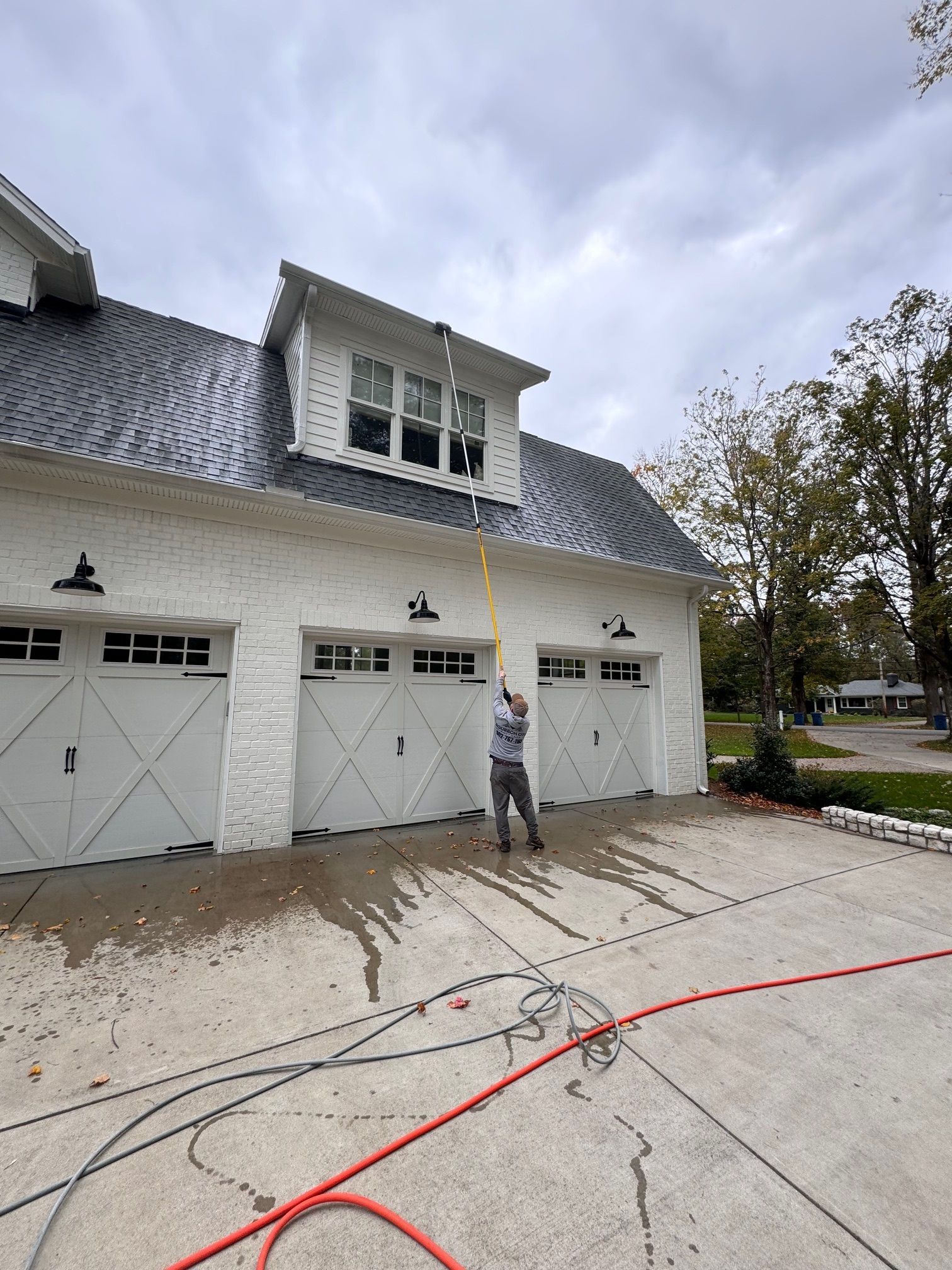 Person cleaning the gutters on a white brick building with a black roof using a long yellow tool. Overcast sky.