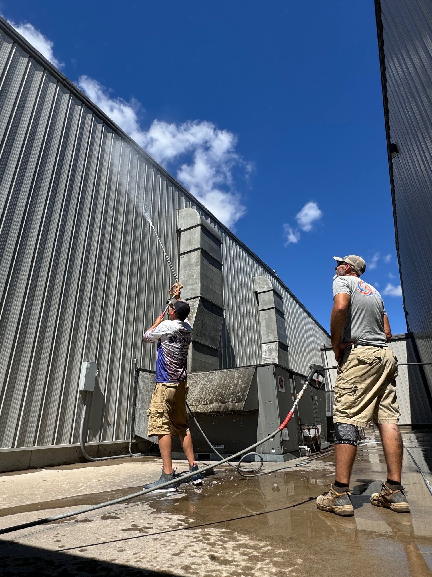 Two people pressure washing a building's exterior on a sunny day.