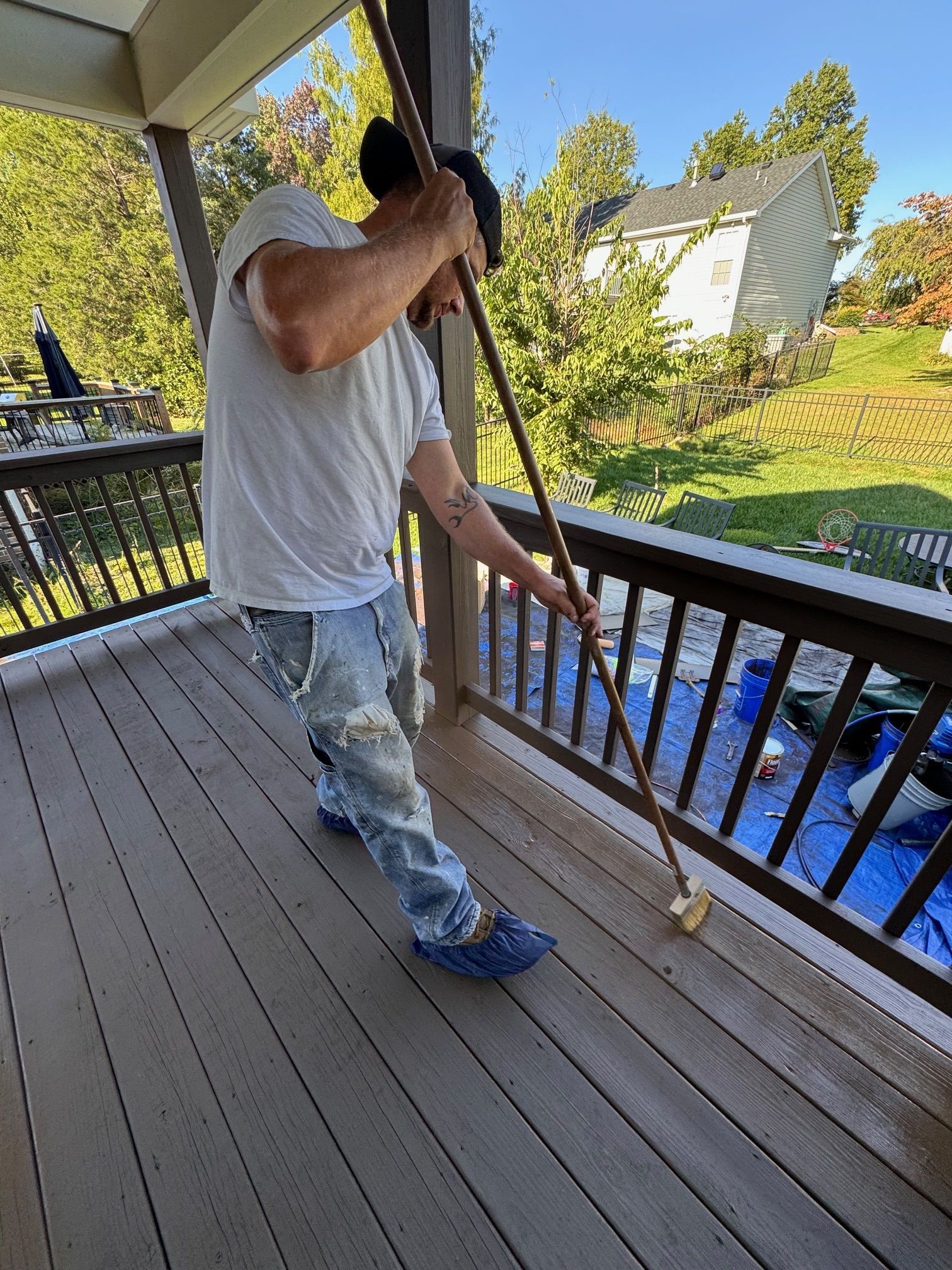Man scrubbing a wooden deck with a long-handled brush on a sunny day.