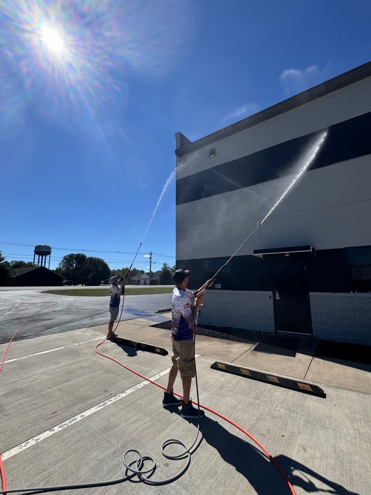 Two people pressure washing a building with bright sun overhead.