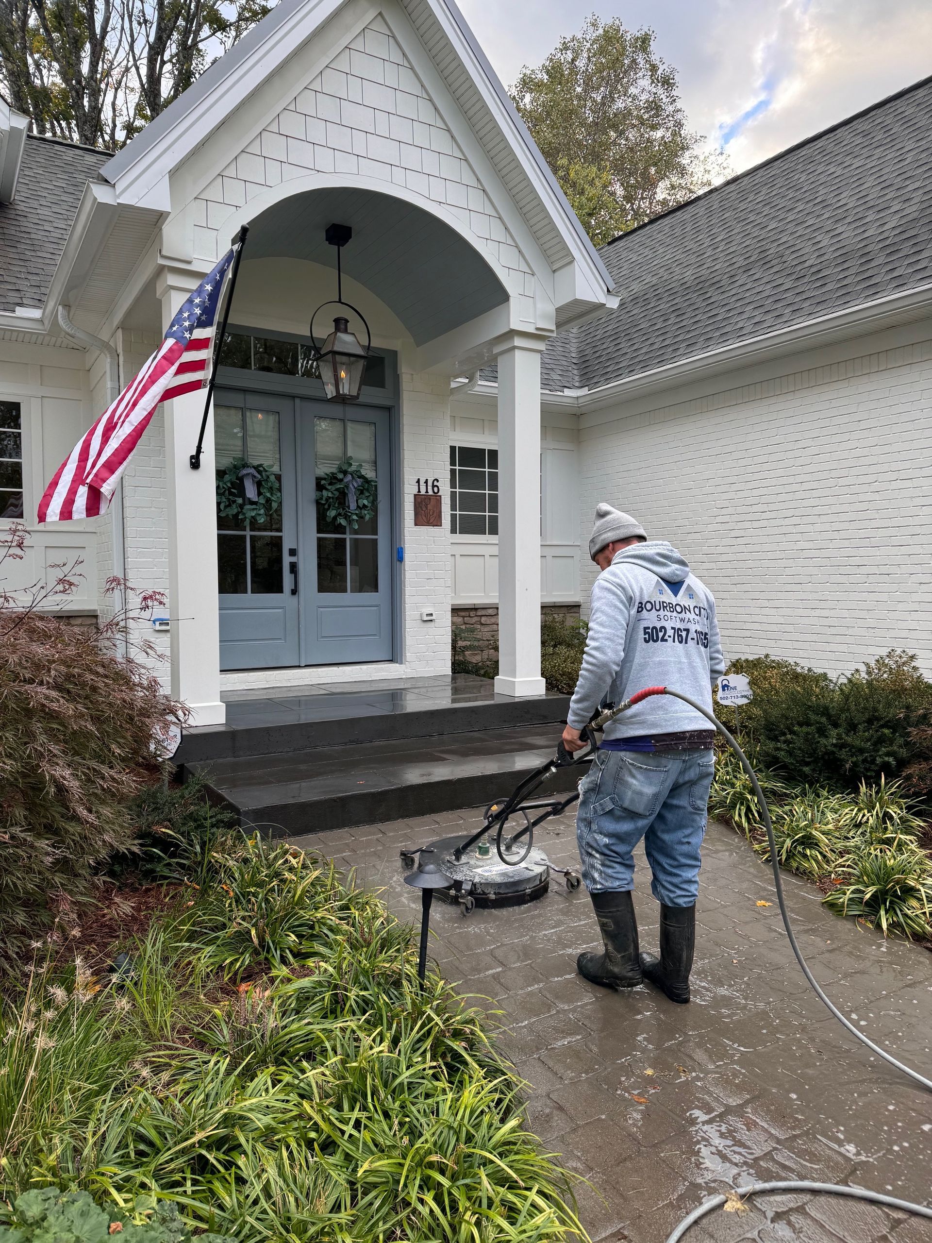 Man pressure washing a concrete walkway in front of a white house with a blue door and American flag.