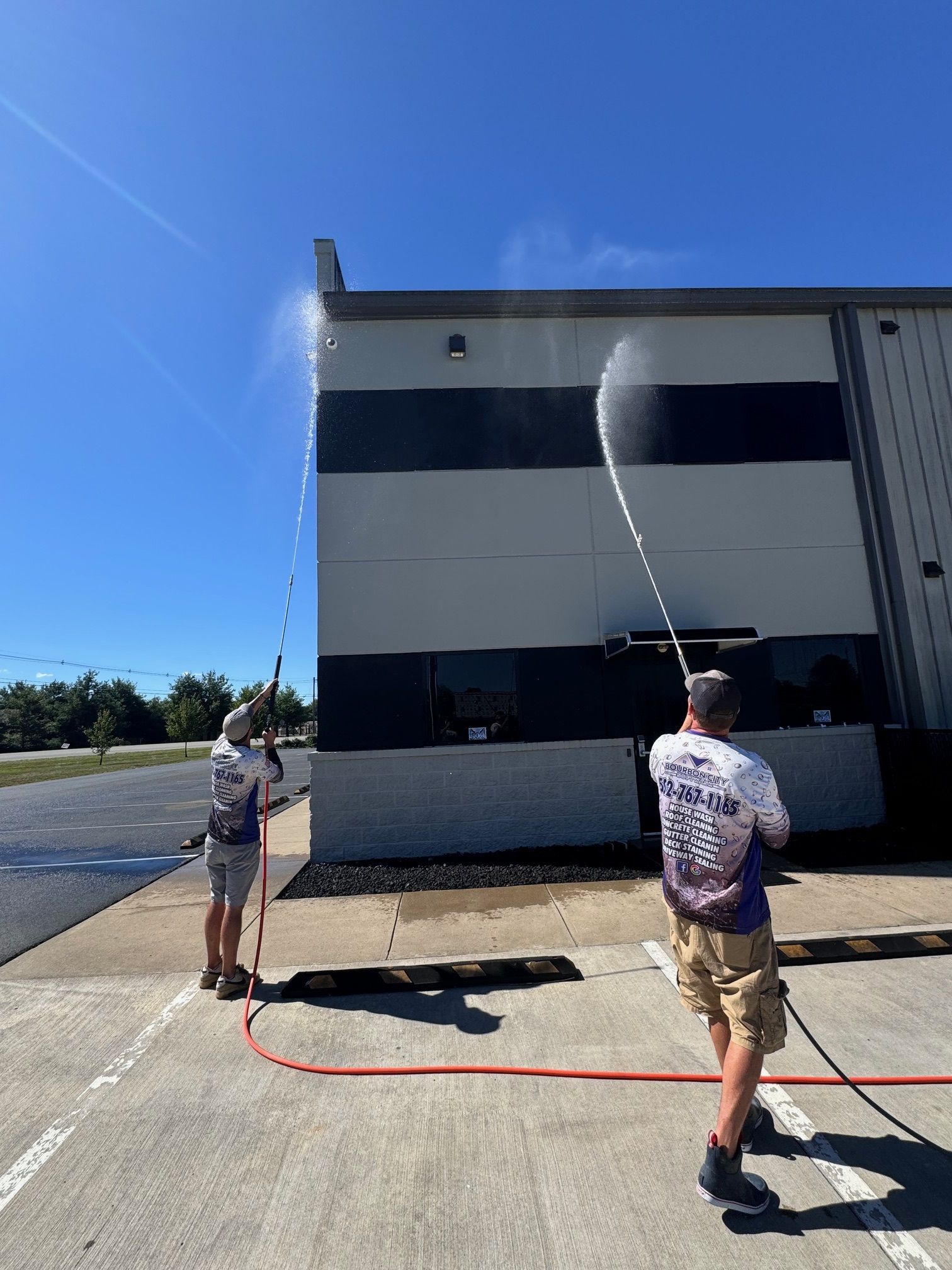 Two people pressure washing a building's exterior on a sunny day.