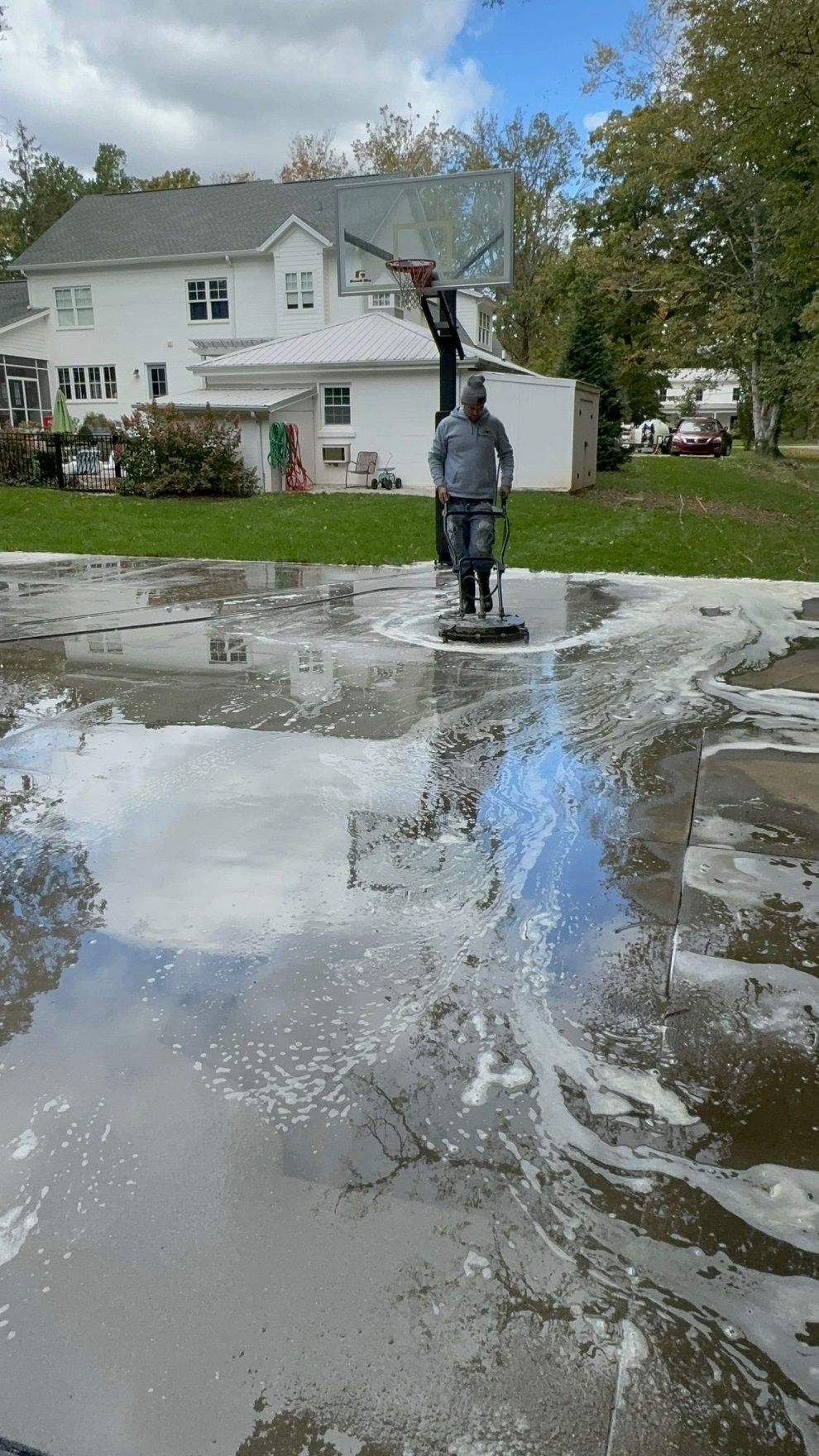 Person pressure washing a concrete surface. Foam and water spray. House, trees, basketball hoop visible.