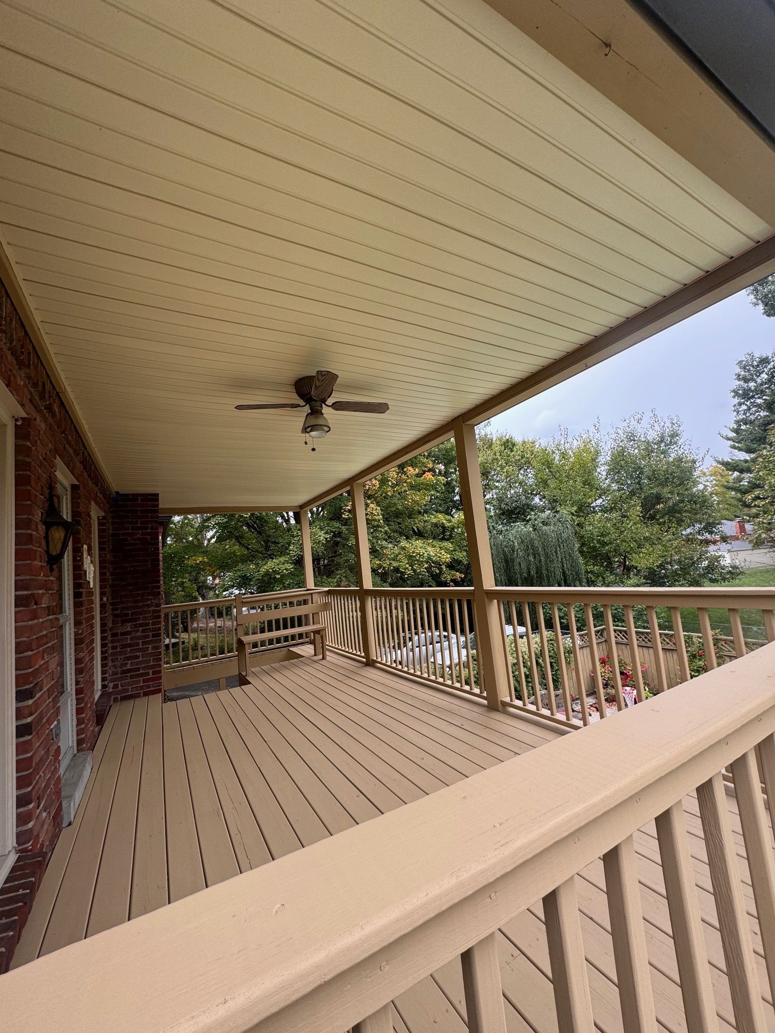 Covered porch with wood deck, ceiling fan, and railing. Trees visible in the background.