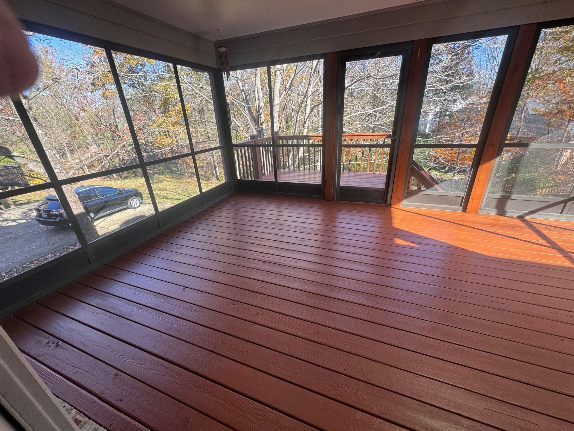 Screened porch with reddish-brown wood floor and glass windows overlooking trees and a car.