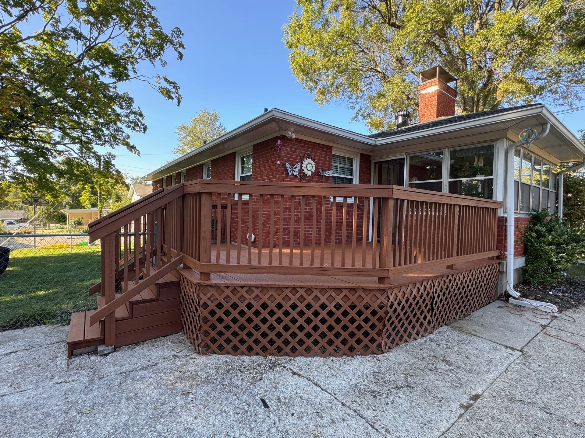 Red brick house with brown wooden deck and lattice skirting, surrounded by a concrete driveway and green grass.