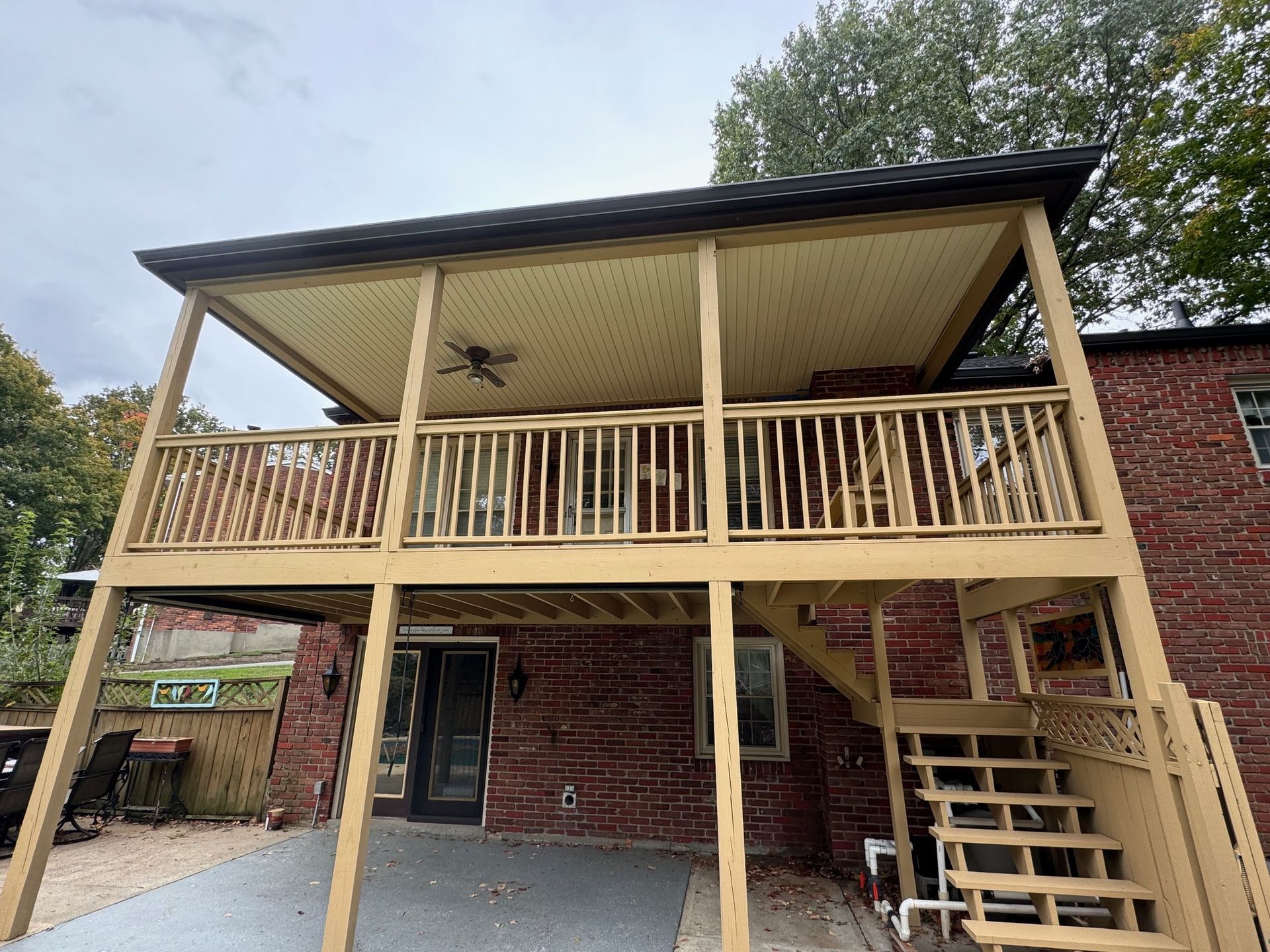 Two-story wooden deck with a covered upper level. Beige ceiling, brown railing, against a brick wall.