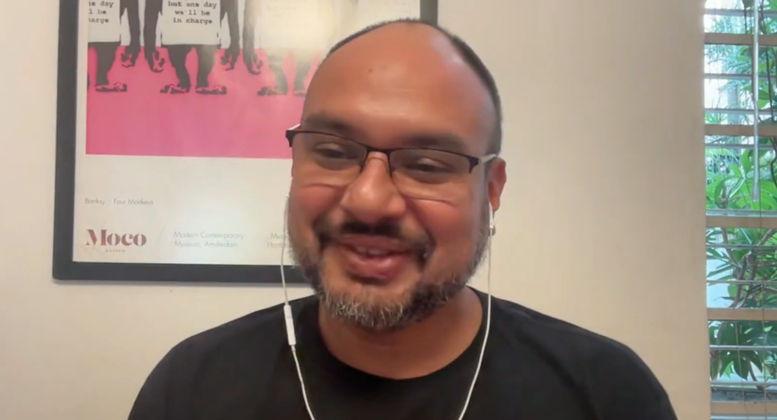Homem de óculos e barba, vestindo camisa preta, olhando para a câmera e sorrindo, com fones de ouvido, em ambiente interno.