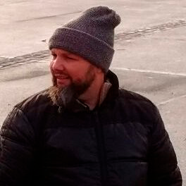 Homem barbudo usando um gorro cinza e jaqueta preta, olhando para o lado.