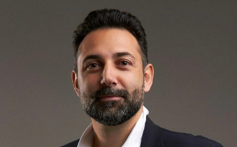 Man with dark hair and beard, wearing a suit jacket and open-collared white shirt, smiling.