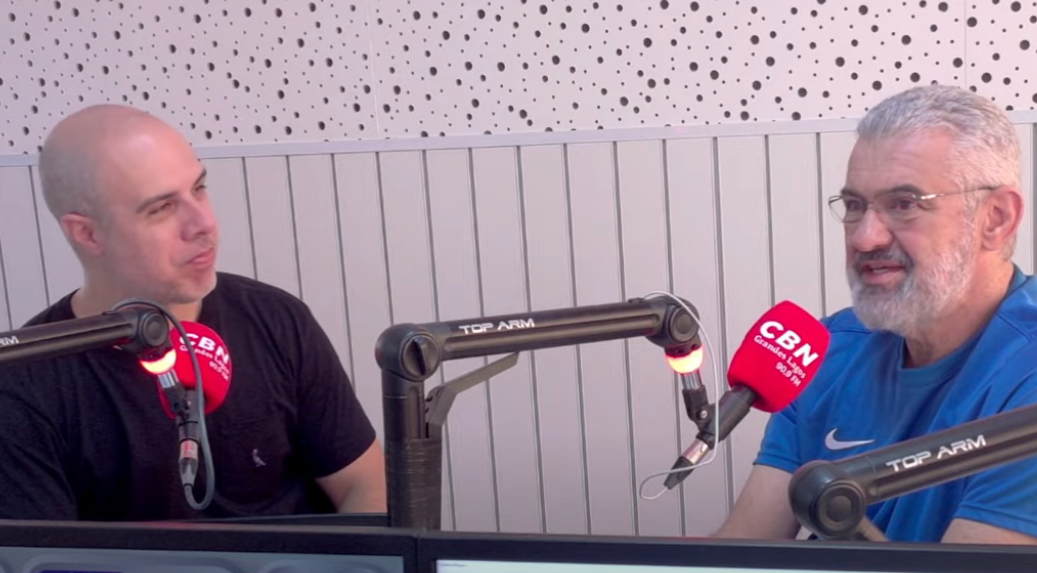 Dois homens sentados em um estúdio de rádio, com microfones à sua frente. Um veste camisa preta, o outro camisa azul e óculos.