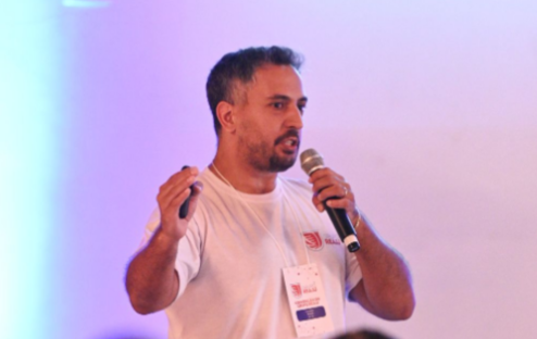 Homem falando ao microfone, segurando um telefone, no palco com um fundo desfocado.