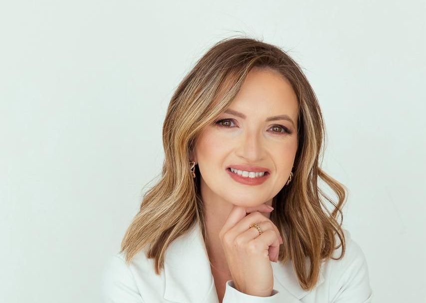 Mulher com cabelos loiros longos e ondulados, sorrindo, mão no queixo, vestindo um blazer branco.