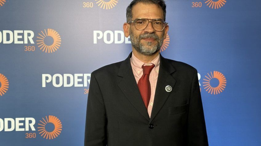 Man in a suit, tie, and glasses stands in front of a blue backdrop with the