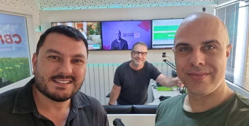 Três homens sorrindo em um estúdio de rádio. O do meio está atrás de uma mesa, os outros na frente, com telas ao fundo.