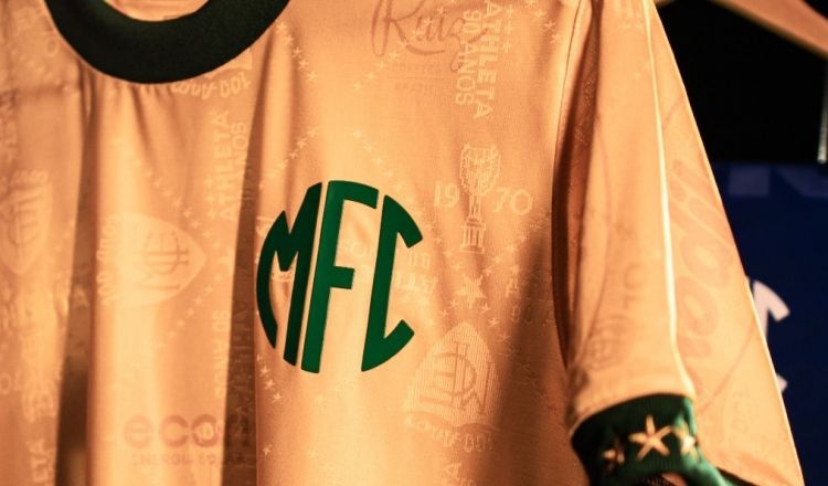 Close-up de uma camisa de futebol dourada com detalhes em verde e um logotipo verde
