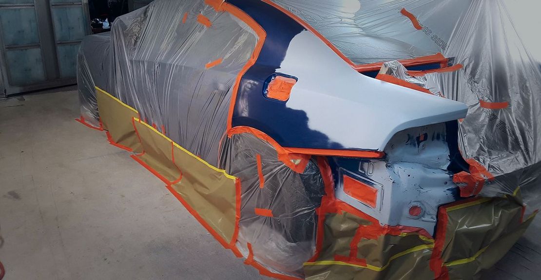 Car partially masked for paint. Blue and white paint is visible, alongside protective plastic and tape in a workshop setting. | Stu's Collision Center
