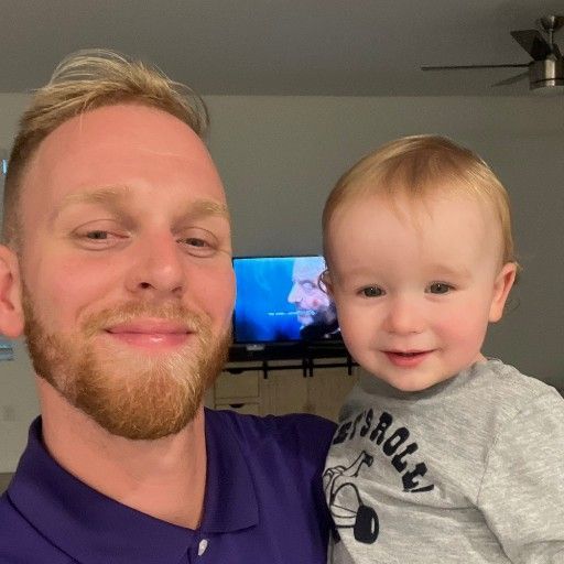 Man with a reddish beard holding a toddler, smiling. They are inside a home.