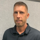 Man with short brown hair and stubble, wearing a black polo shirt, looking at the camera.