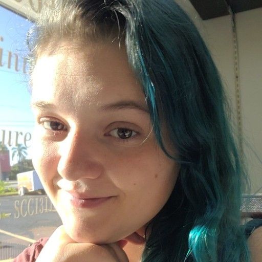 Woman with teal hair smiles, resting chin on hand; sunny outdoor setting with business sign in background.