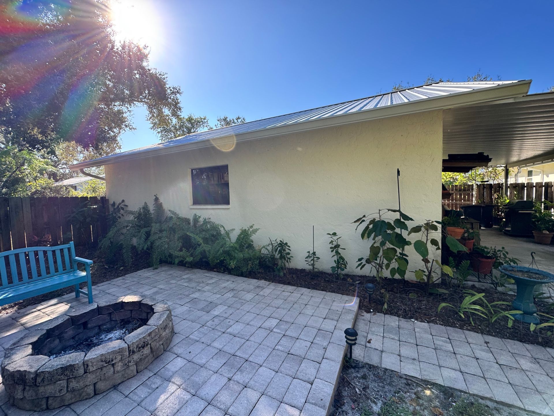 Backyard patio with fire pit, blue bench, and plants. Sunny, clear sky.