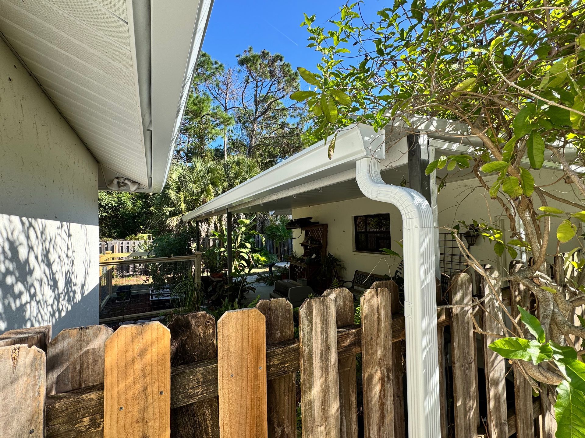 View of a white house exterior with a gutter, wooden fence, and lush greenery under a blue sky.