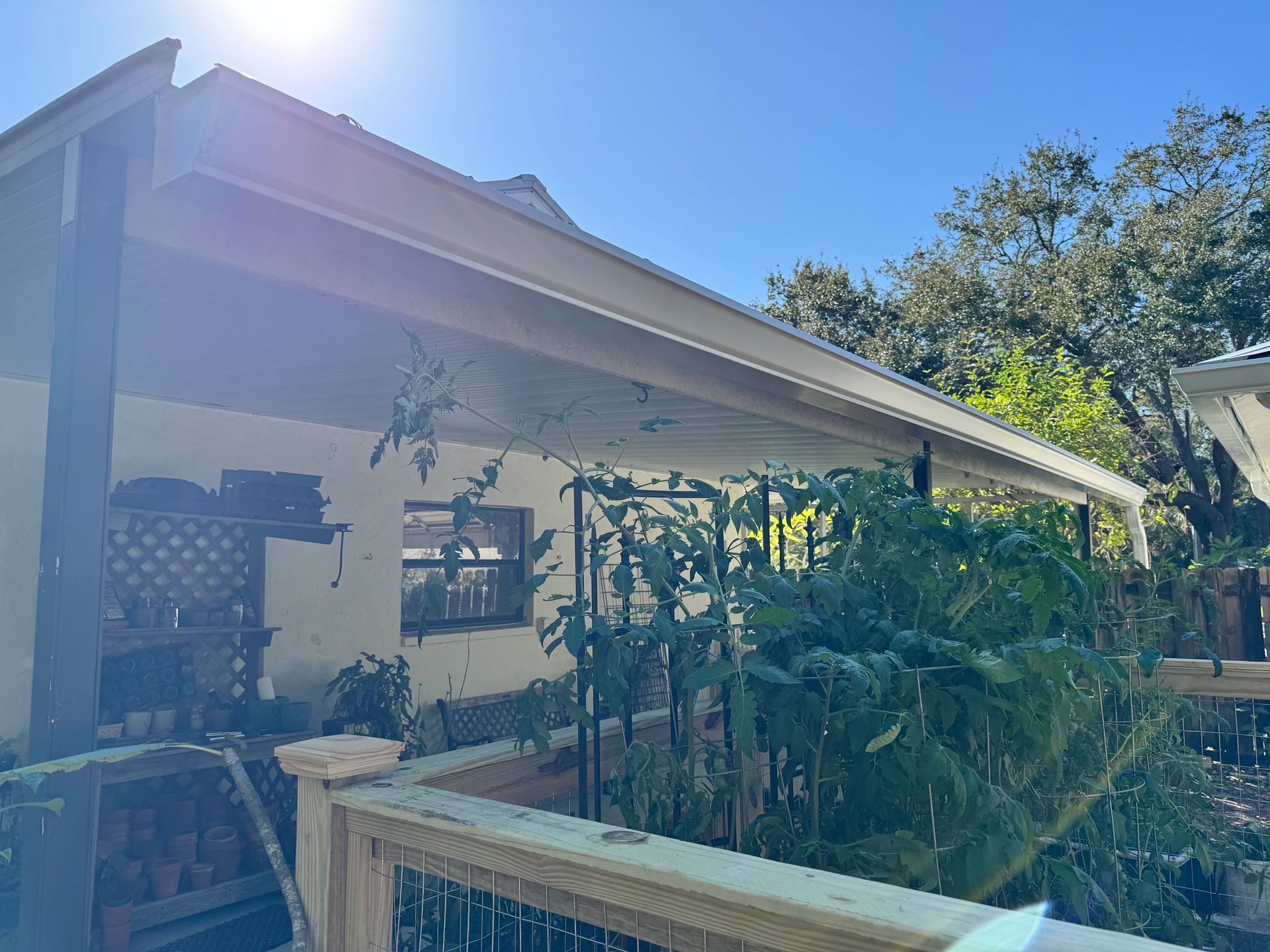 Covered porch with plants in foreground and a wooden fence, under a blue sky.
