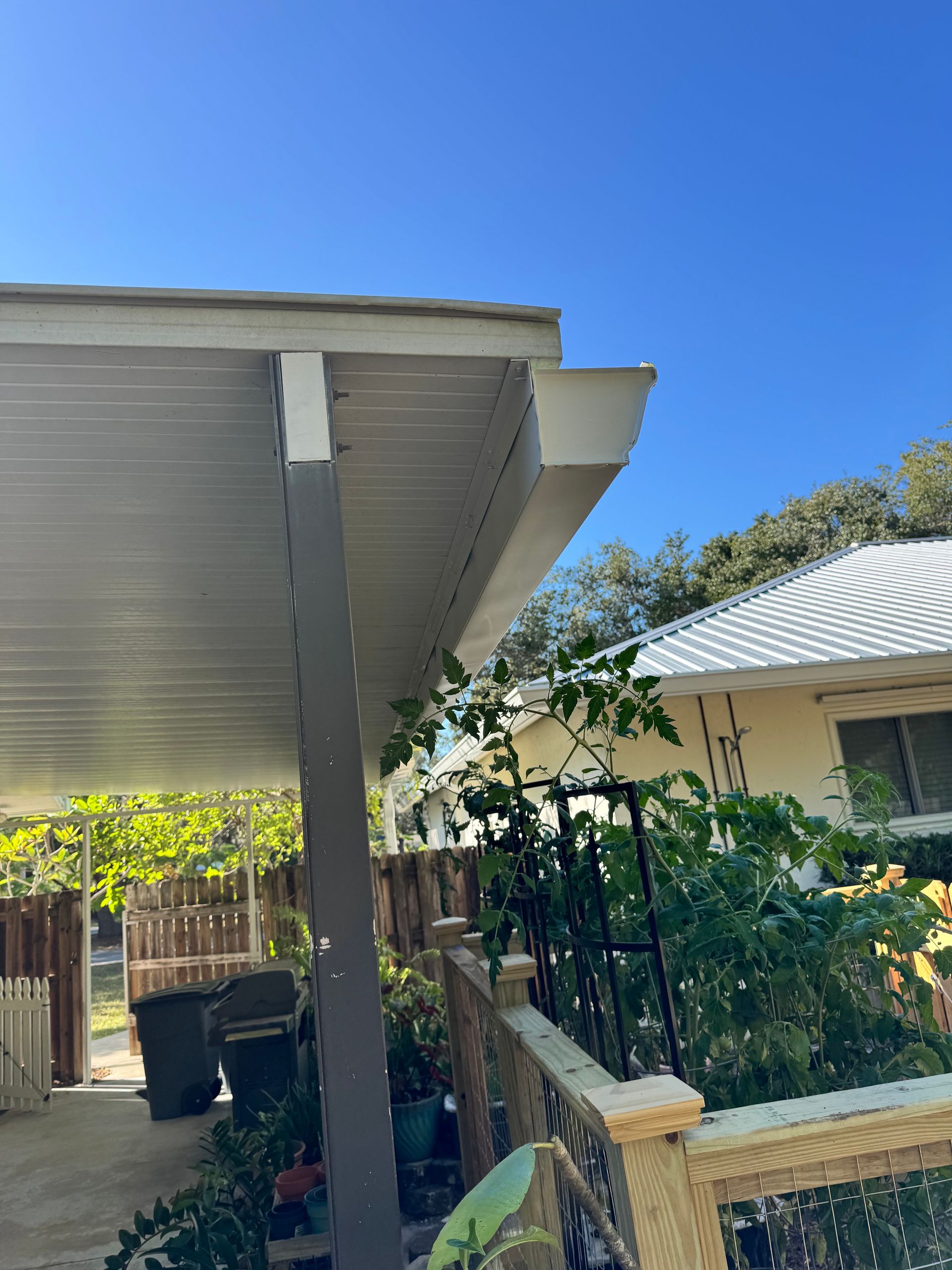 View of a white roof with a gutter, supported by a gray column. Plants and a house are in the background.