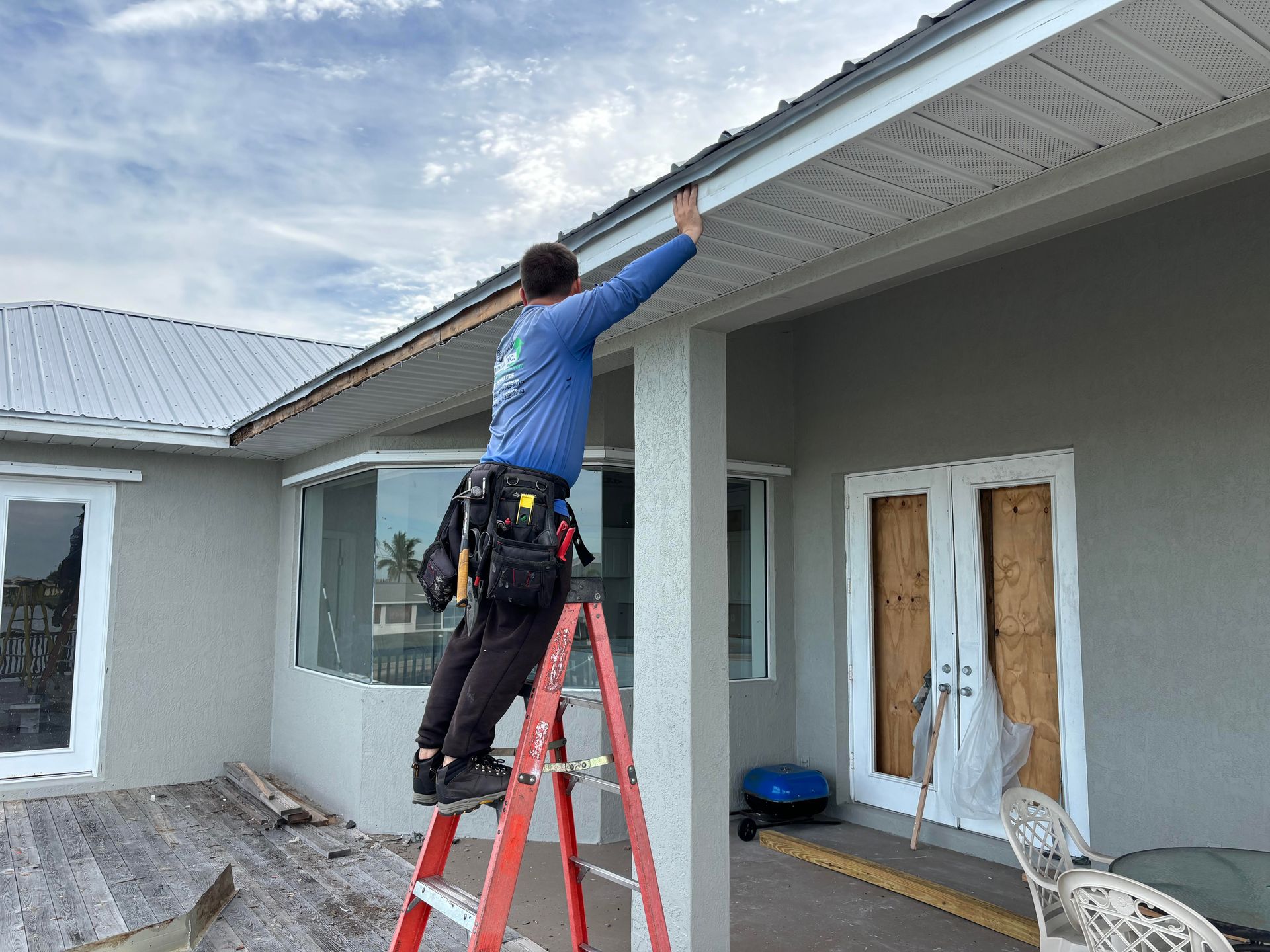 Person on a ladder installing soffit. Gray house, cloudy sky.