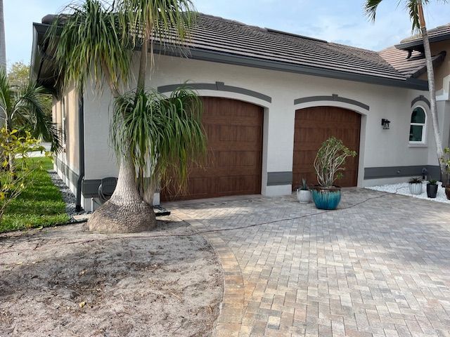 Garage with brown arched doors, light stucco walls, and paved driveway. A palm tree is in the foreground.