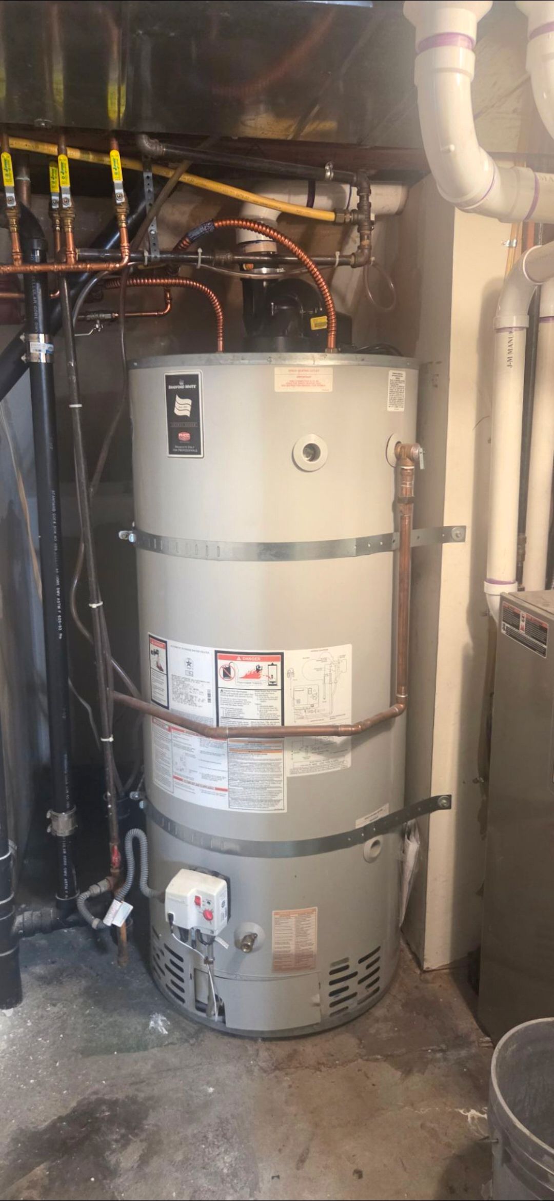 Tall beige water heater in a basement utility area with pipes and wiring around it