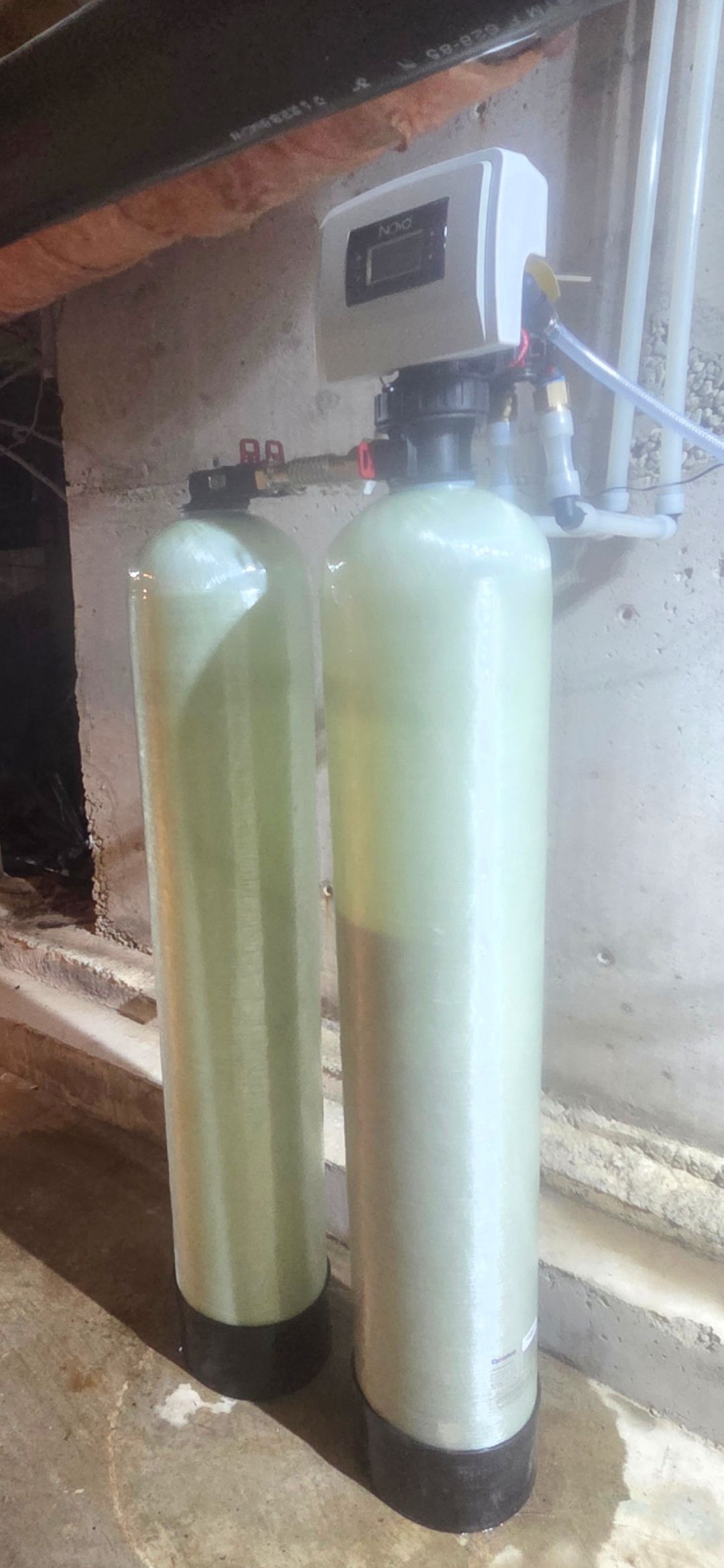 Two tall white water filter tanks connected to a wall-mounted control unit in a utility area