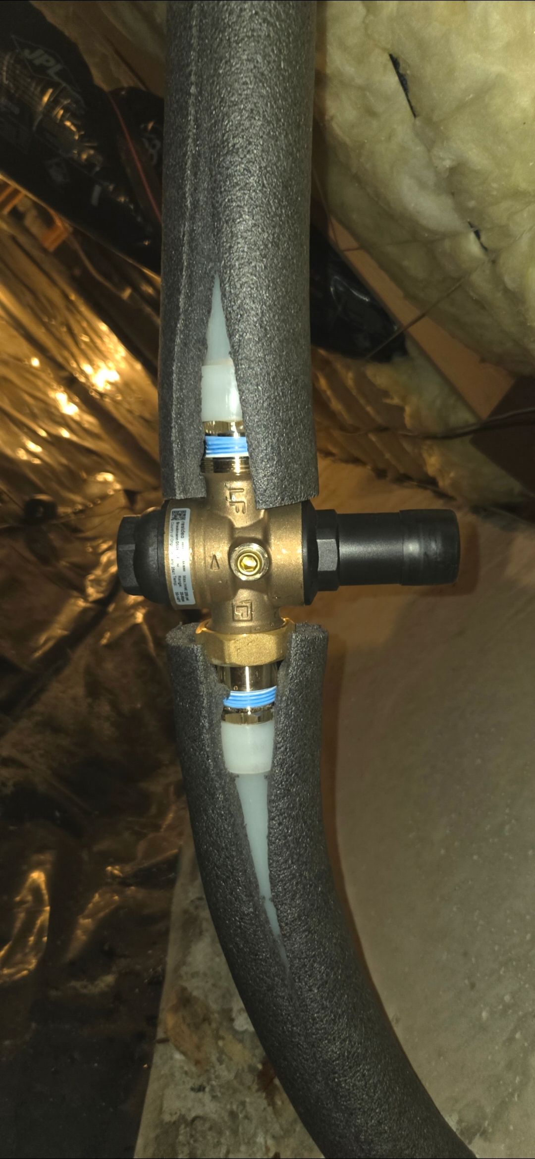 Brass valve on black insulated pipe in an attic with exposed insulation and wood framing