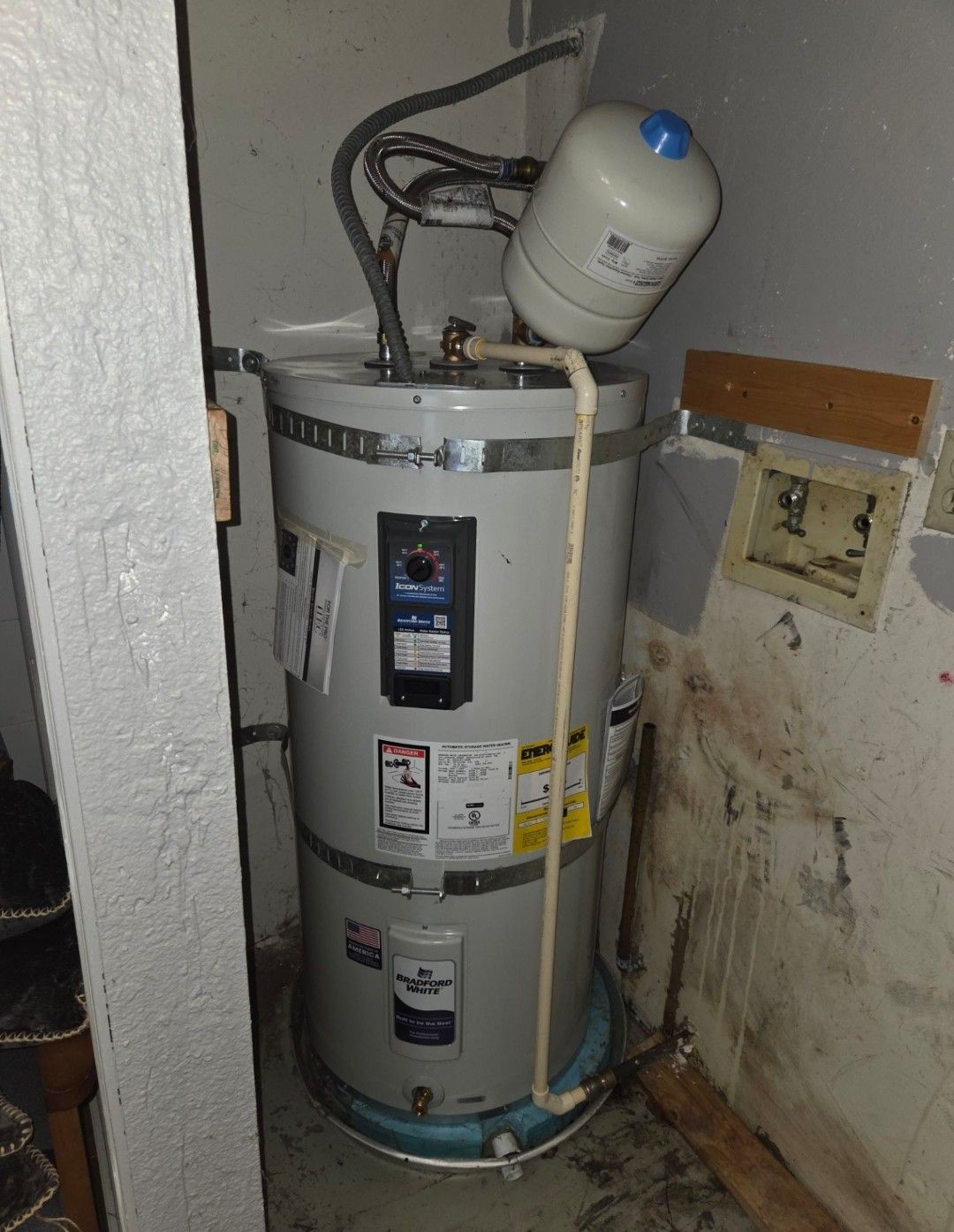 White water heater in a narrow utility closet with pipes and a small tank on top, beside a stained wall.