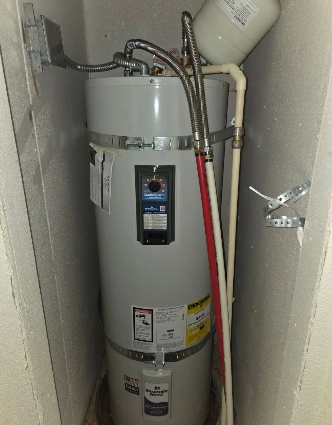 Water heater in a narrow utility closet with pipes, valves, and a vent pipe.