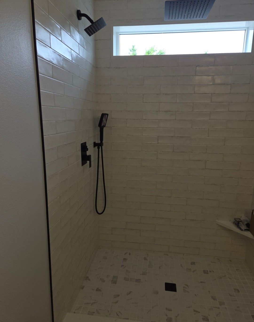 Empty white-tiled shower with black fixtures, pebble floor, and a small frosted window near the ceiling