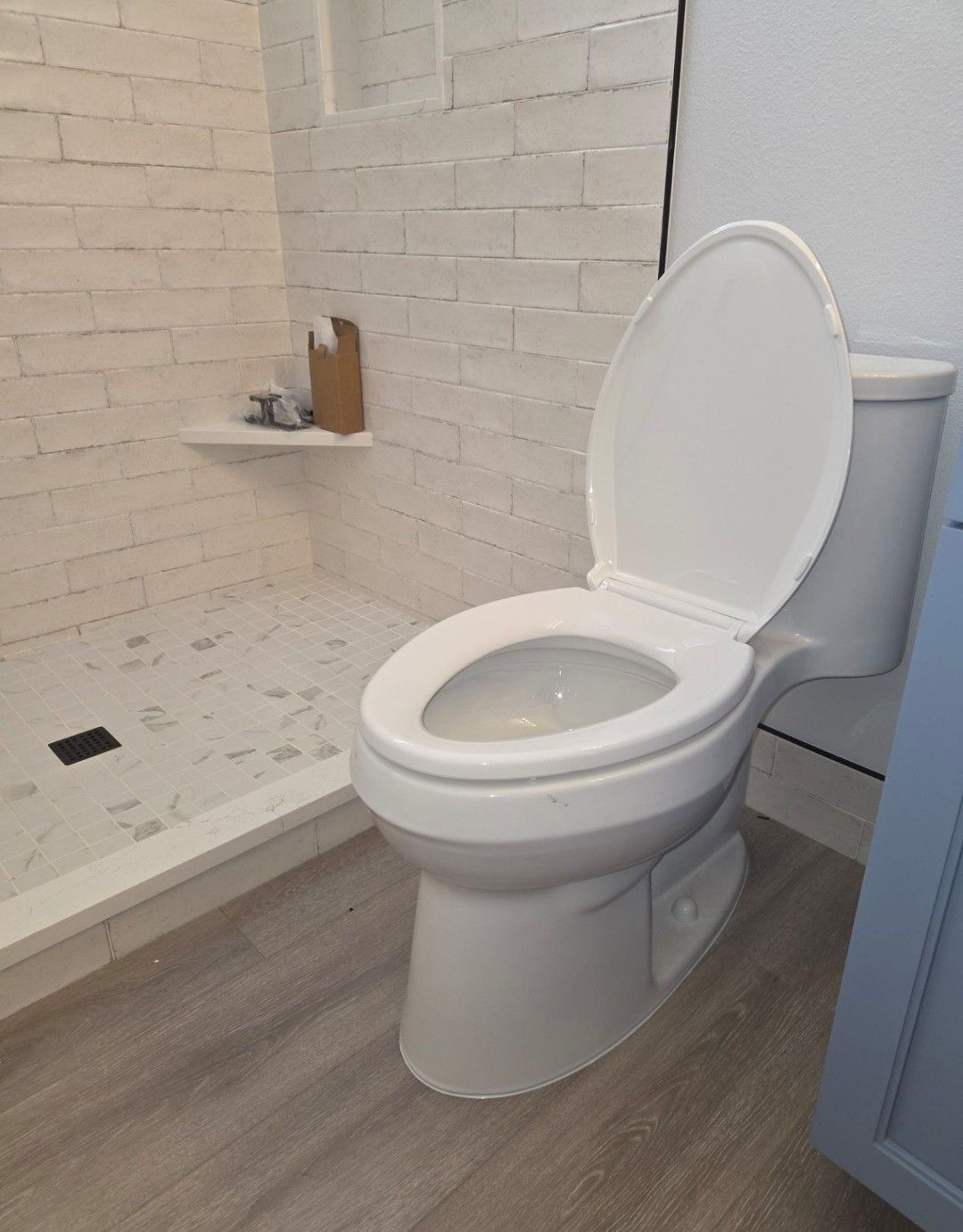 White toilet with lid up in a tiled bathroom with a small shelf and wood-look floor.