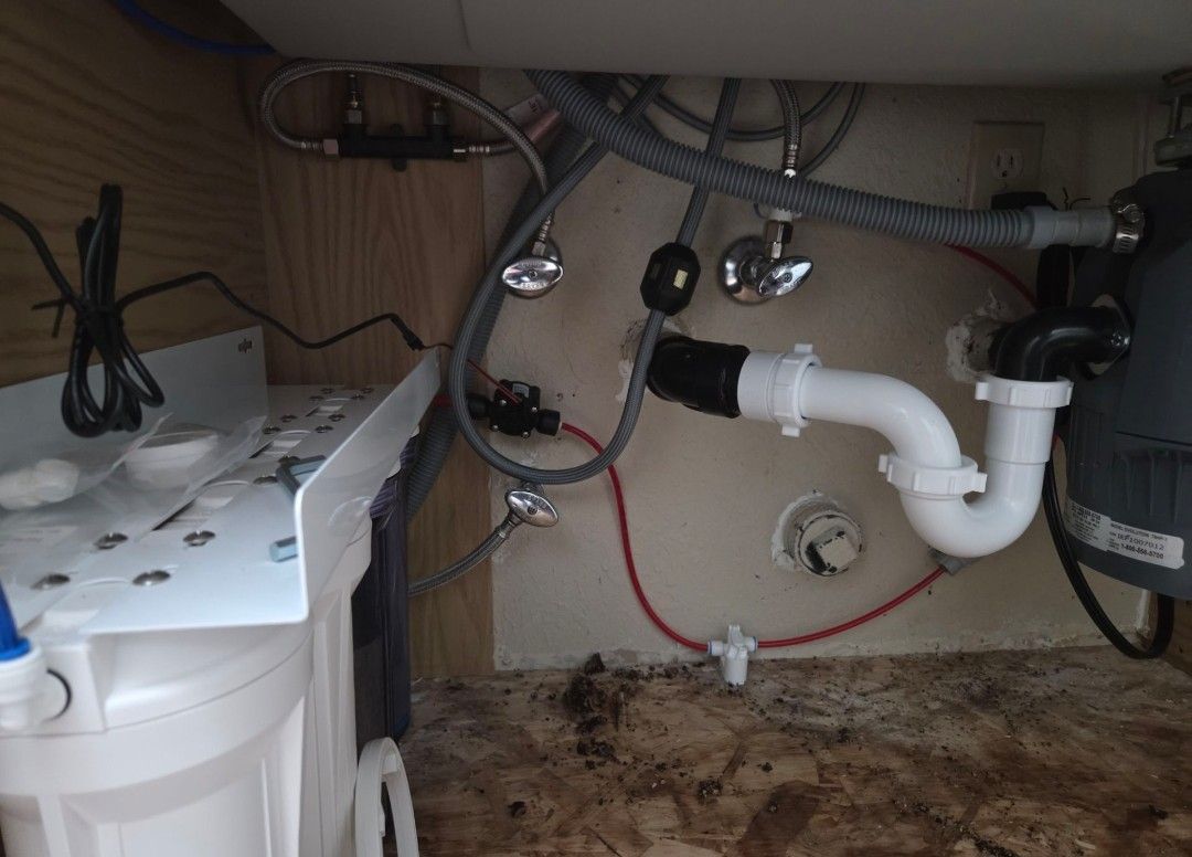 Under-sink plumbing and drainage pipes with a white appliance at left and a wet, dirty cabinet floor