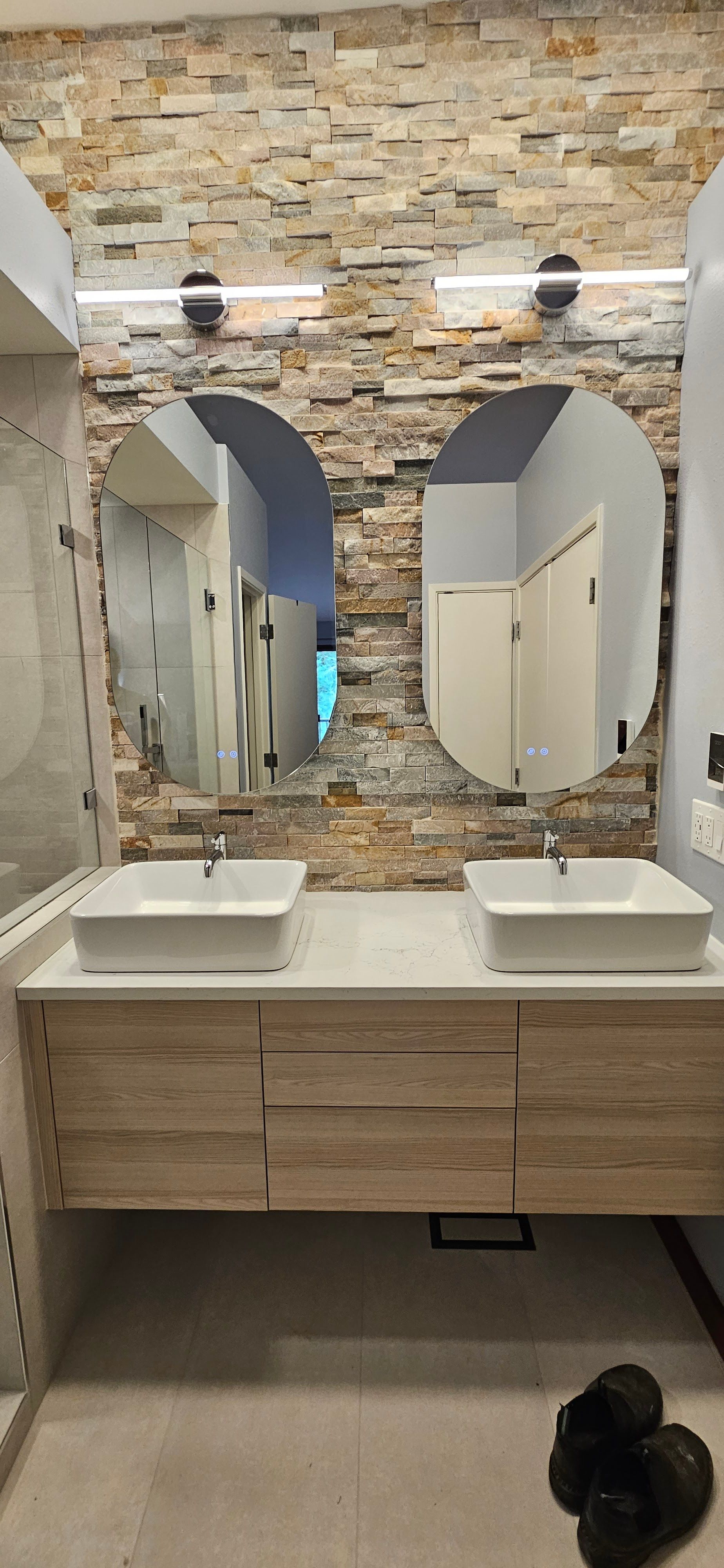 A double vanity with white square sinks, light wood cabinets, and two oval mirrors mounted on a textured stone wall.