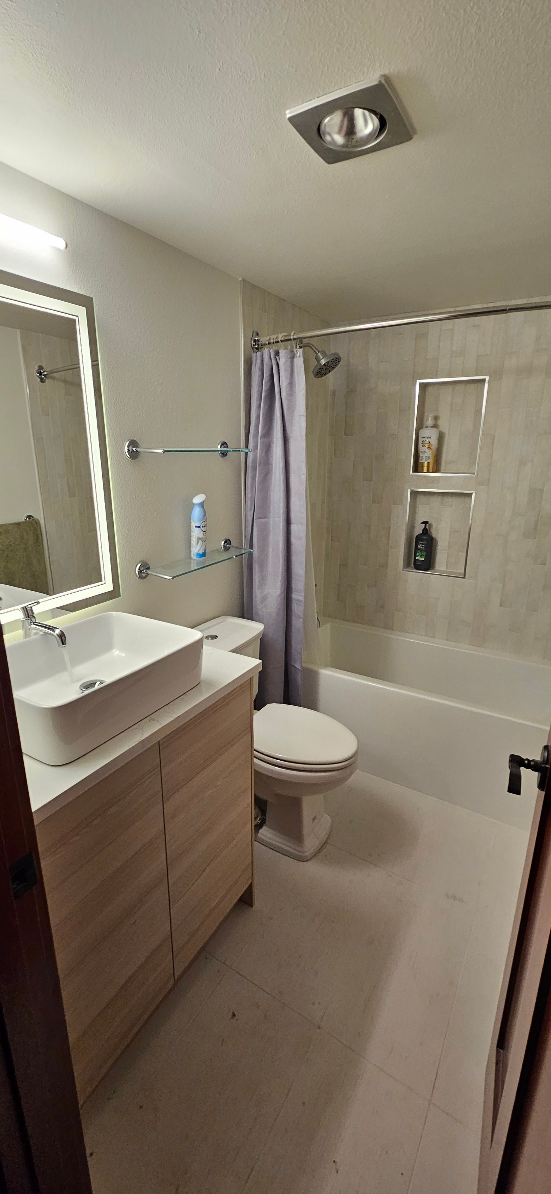 A modern bathroom with a light wood vanity, vessel sink, illuminated mirror, a toilet, and a shower-tub with glass niches.