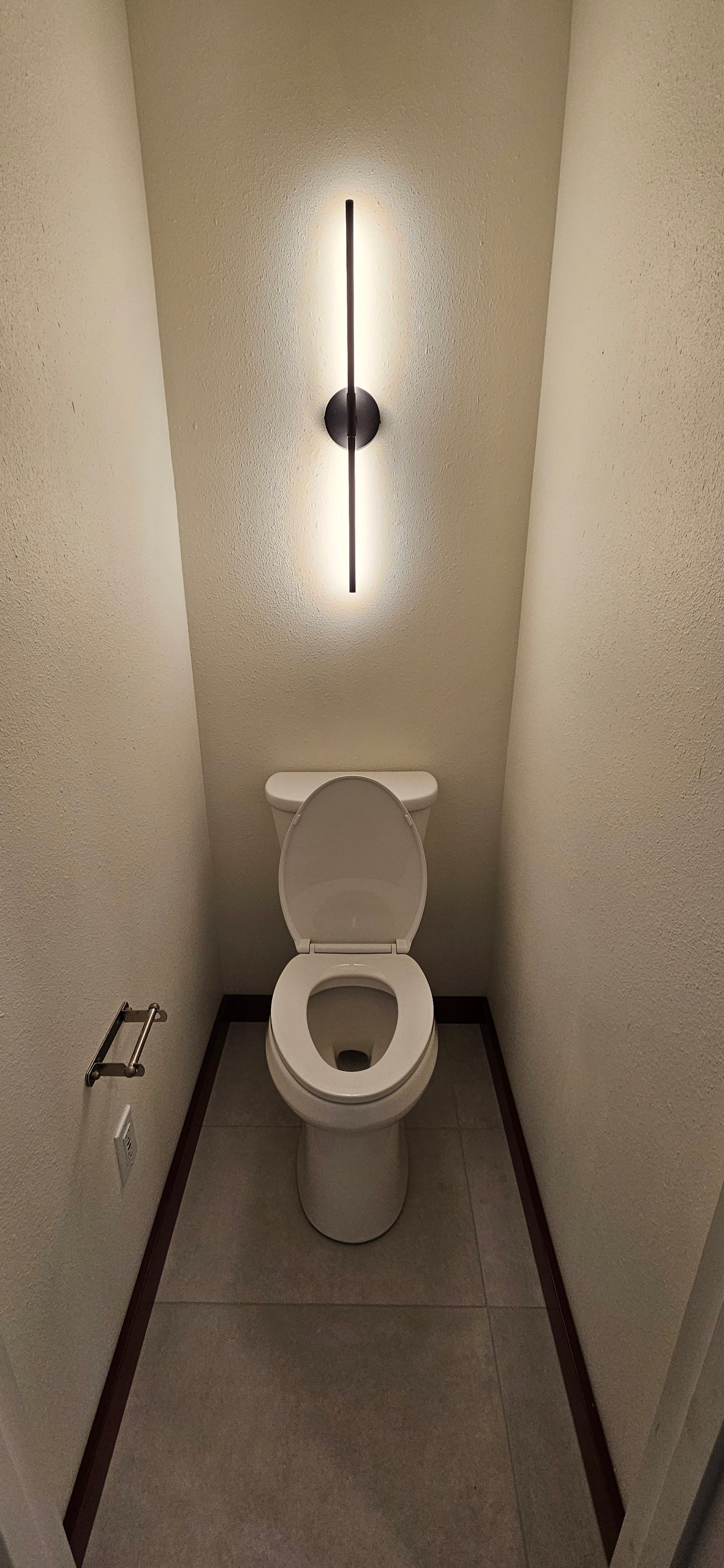 A small, narrow restroom featuring a white toilet, gray tiled floor, light textured walls, and a modern vertical wall light.